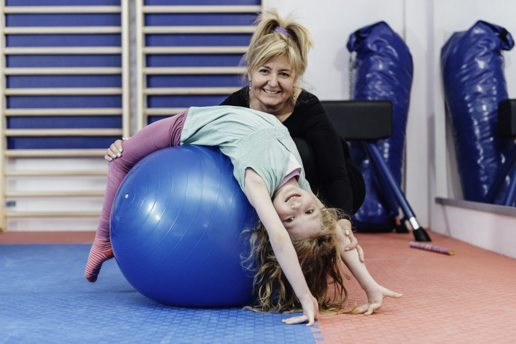 Physical therapist working with cute preschooler little girl in gymnasium, stretching over fitness ball