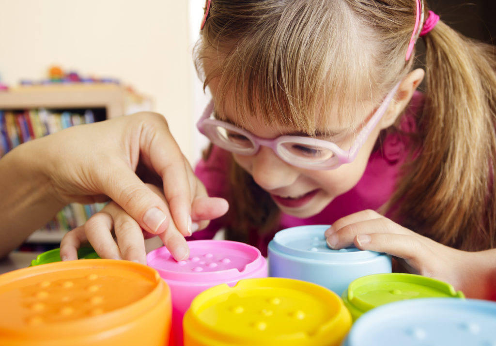 Smiling child with disability touching textured cups with her teacher Little girl with poor vision is playing with colorful tactile toy cups as a part of occupational therapy with her teacher.